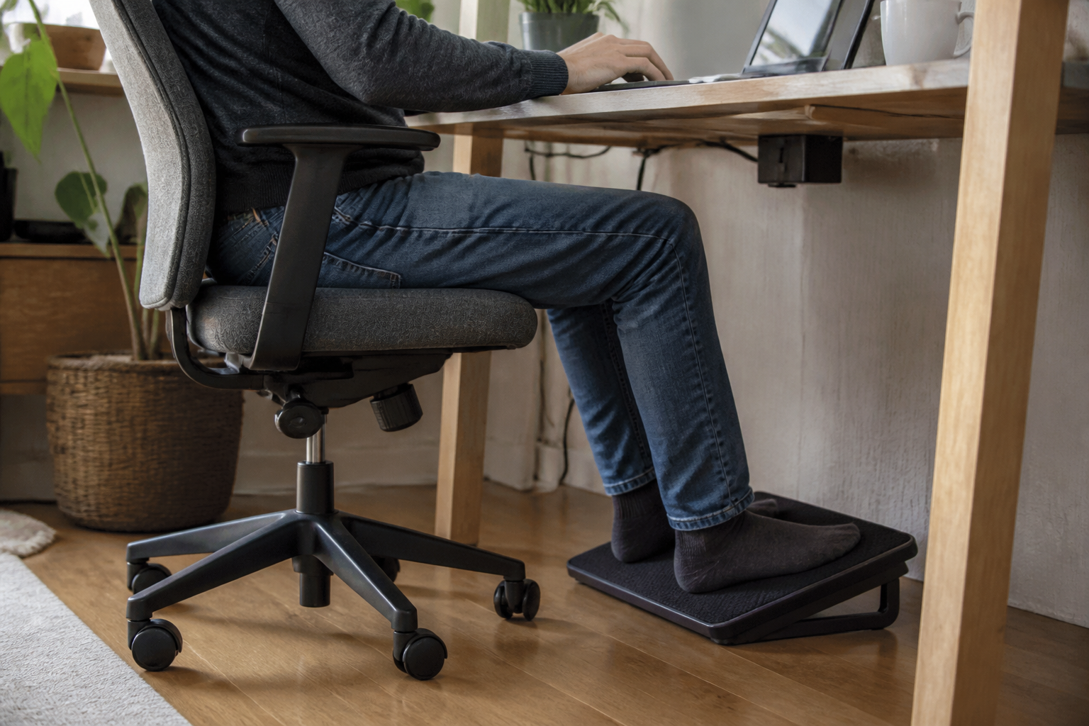 A seated home-office worker has both feet supported on a simple angled footrest beneath a fixed-height desk.